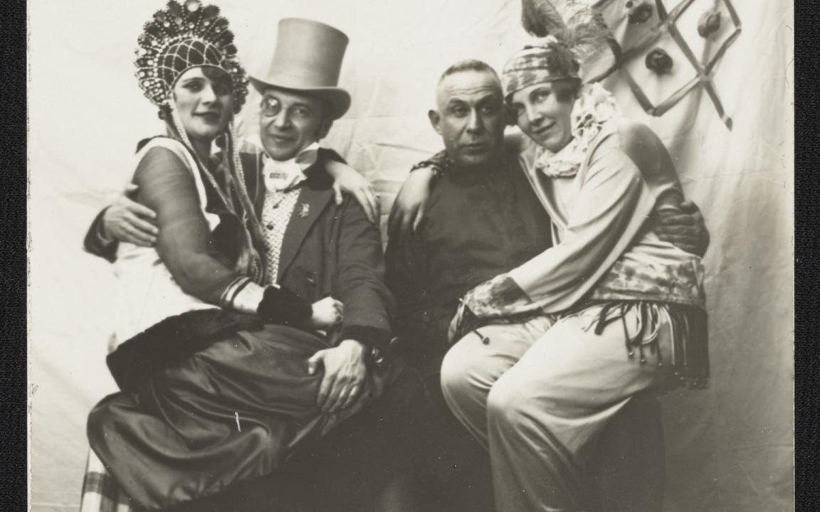 Photo of two women sitting on the laps of two men, all four wearing elaborate clothes with various headdresses