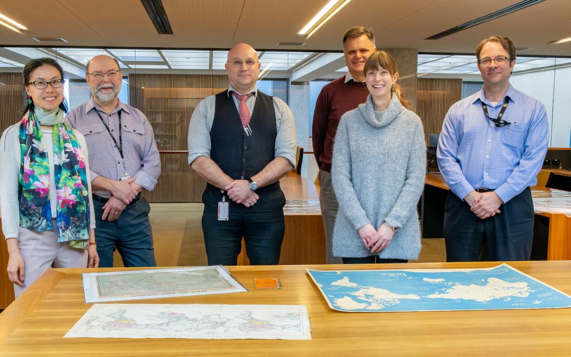 Two women and four men, who make up the Library's Special Collections Research & Support team, standing and smiling in the Special Collections Reading Room in front of a table with three maps on it