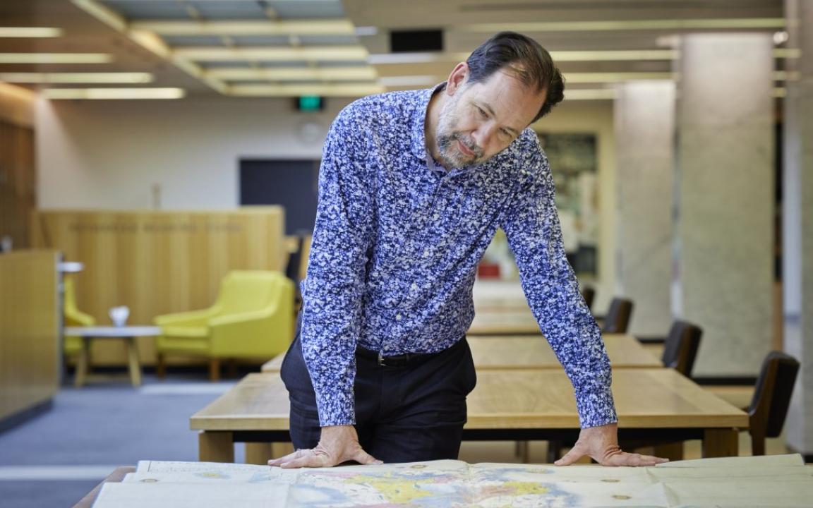 Man with brown hair and a grey and brown beard sitting at a table with a large map spread over it