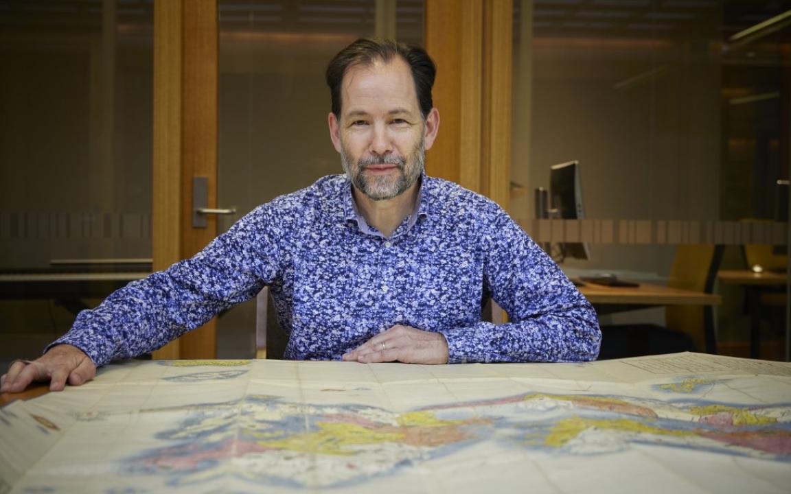 Man with brown hair and a grey and brown beard sitting at a table with a large map spread over it