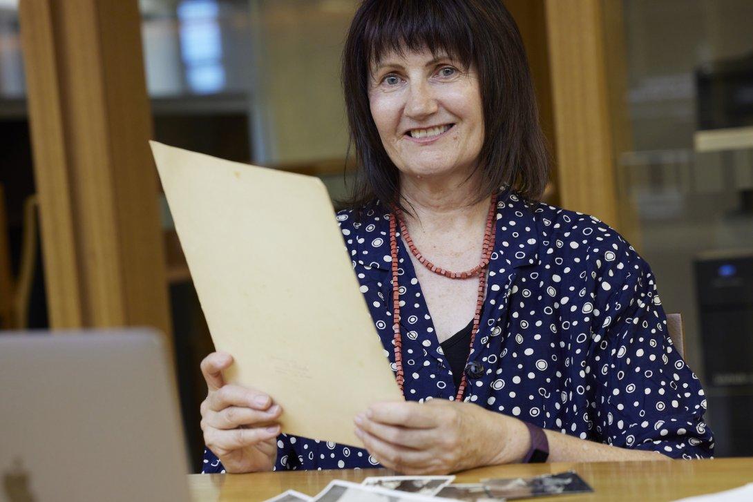 Smiling woman with short black hair and a fringe sitting at a table holding up a piece of paper