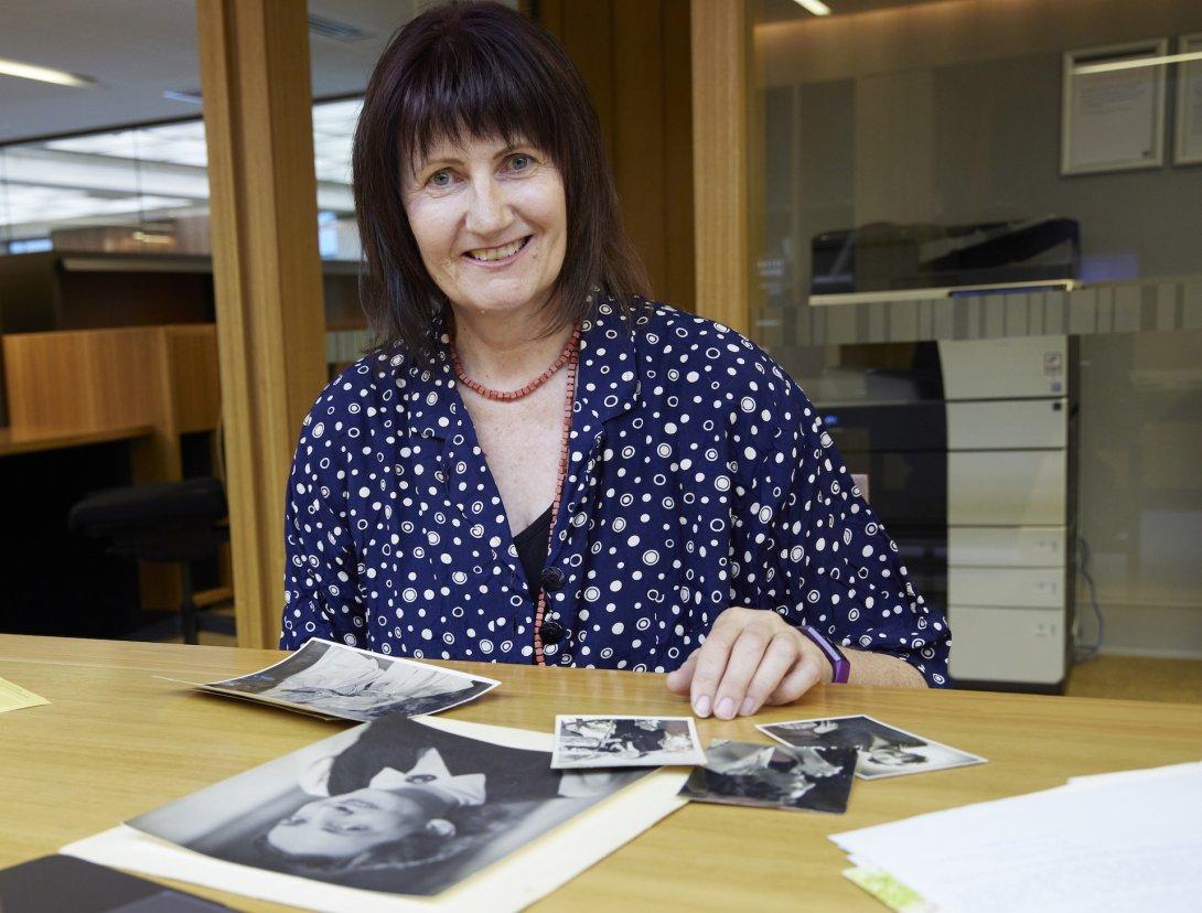 Smiling woman with short black hair and a fringe sitting at a table where several black and white photos of a woman are spread out