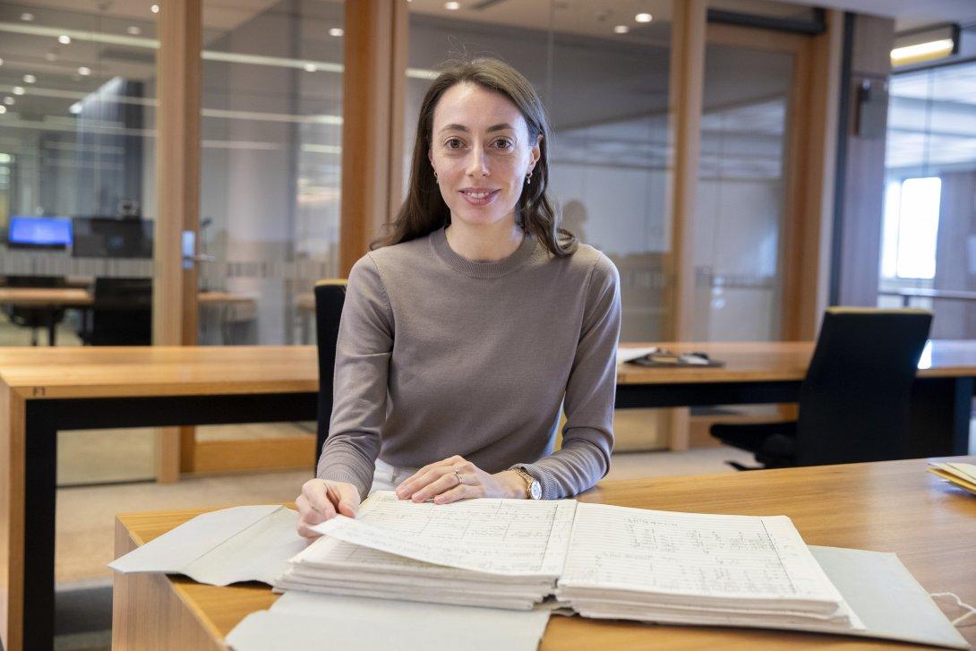 Woman with brown hair and eyes, wearing a brown sweater, sitting at a desk in the Special Collection Reading Room, smiling at the camera. On the desk in front of her is an old book of music and other sheets of paper