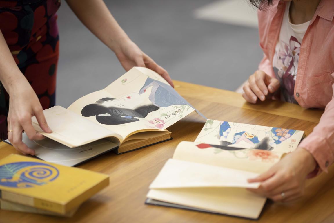 Two books open on a table with people flicking through them and looking at the books' colourful artwork showing drawings of women