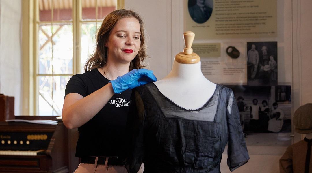 A person stands in Claremont Museum tending to a dress on a mannequin. There is a piano-style instrument, signage and a window in the background.