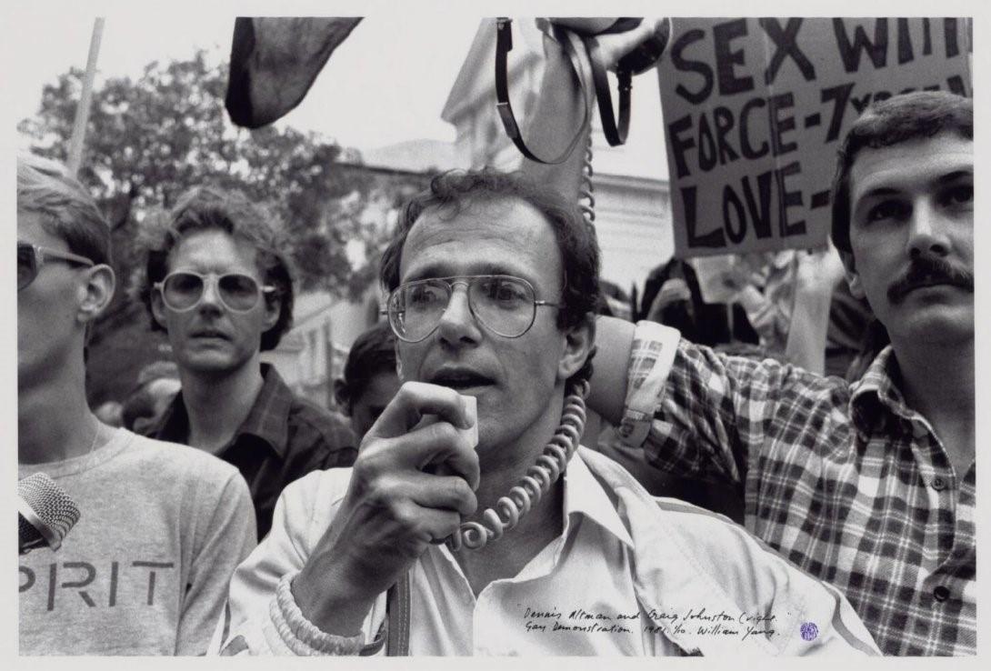 A black and white image of a group of mean demonstrating. The man front and centre is holding a microphone to his mouth, while the man to the right is holding a megaphone speaker up in the air. There are placards and flags in the air in the background.