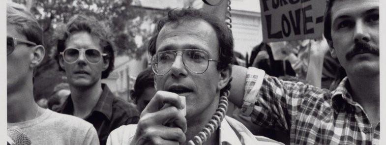 A black and white image of a group of mean demonstrating. The man front and centre is holding a microphone to his mouth, while the man to the right is holding a megaphone speaker up in the air. There are placards and flags in the air in the background.