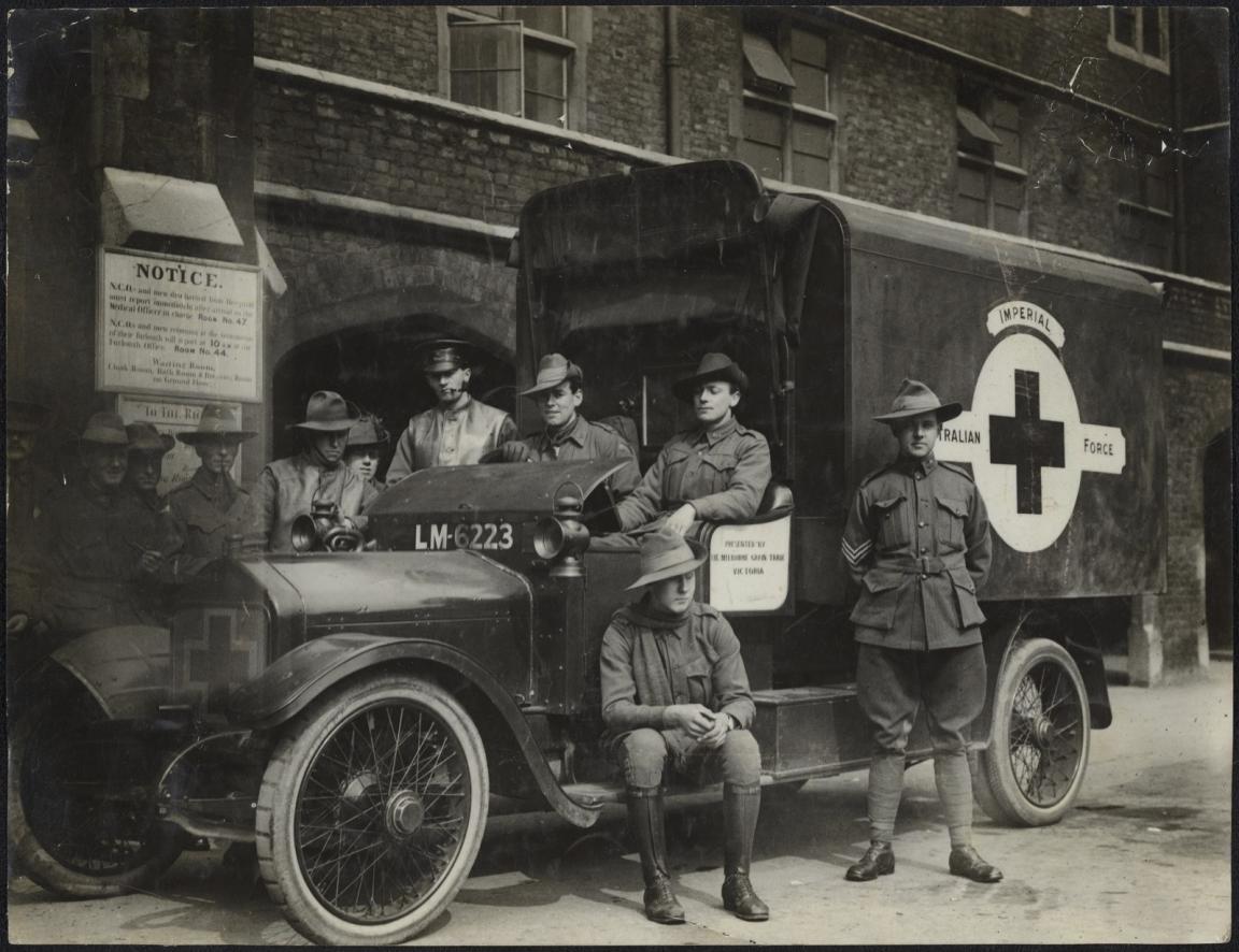 A group of soldiers wearing slouch hats and dressed in uniform are gathered around an Australian Infantry Force ambulance with a Red Cross emblem on the side. Some are seated in the front of the vehicle, while others stand beside it, posing for the photo. The ambulance has a visible license plate "LM-6223." A sign with information about hospital procedures and waiting rooms is posted on the brick wall in the background.