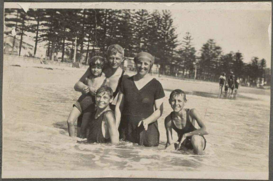 A black-and-white photo of a smiling family in swimsuits, standing together in shallow water at Manly Beach. The background shows pine trees and other beachgoers. The picture was taken in 1923 in New South Wales, Australia.