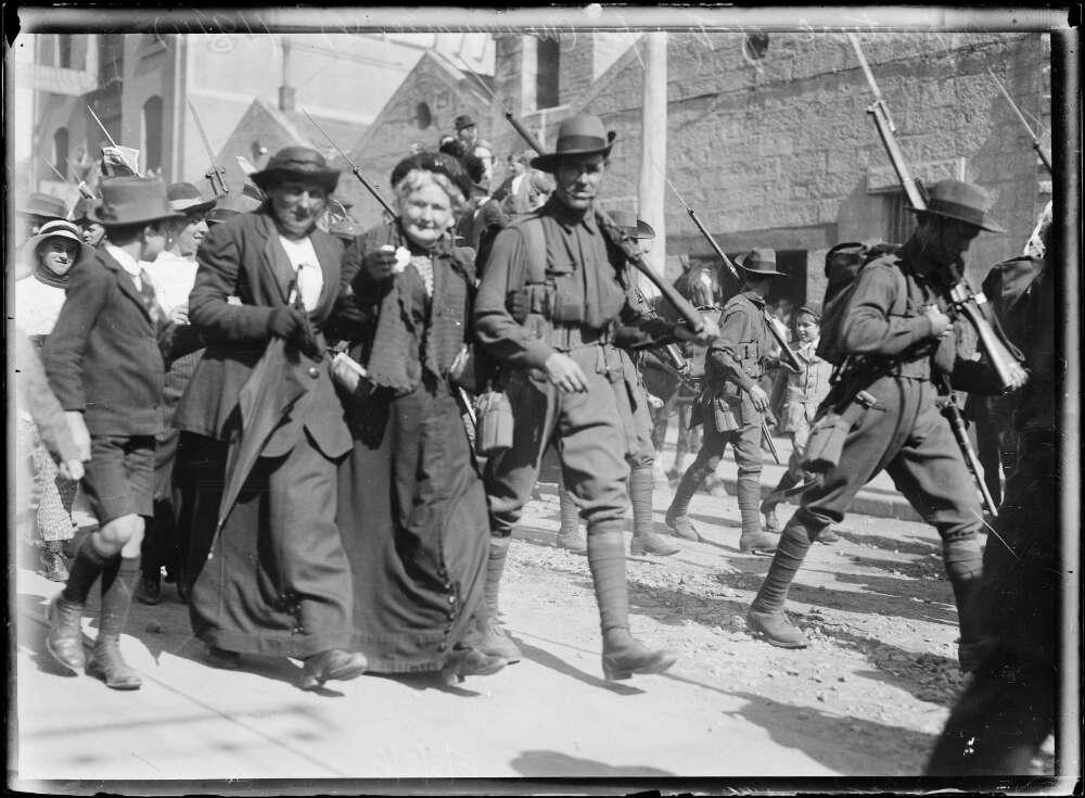 A black-and-white photograph shows a group of soldiers marching down a street with rifles over their shoulders. They are accompanied by civilians, who walk arm-in-arm with the soldiers, smiling and engaging with the crowd. The street is busy with people, and buildings can be seen in the background.