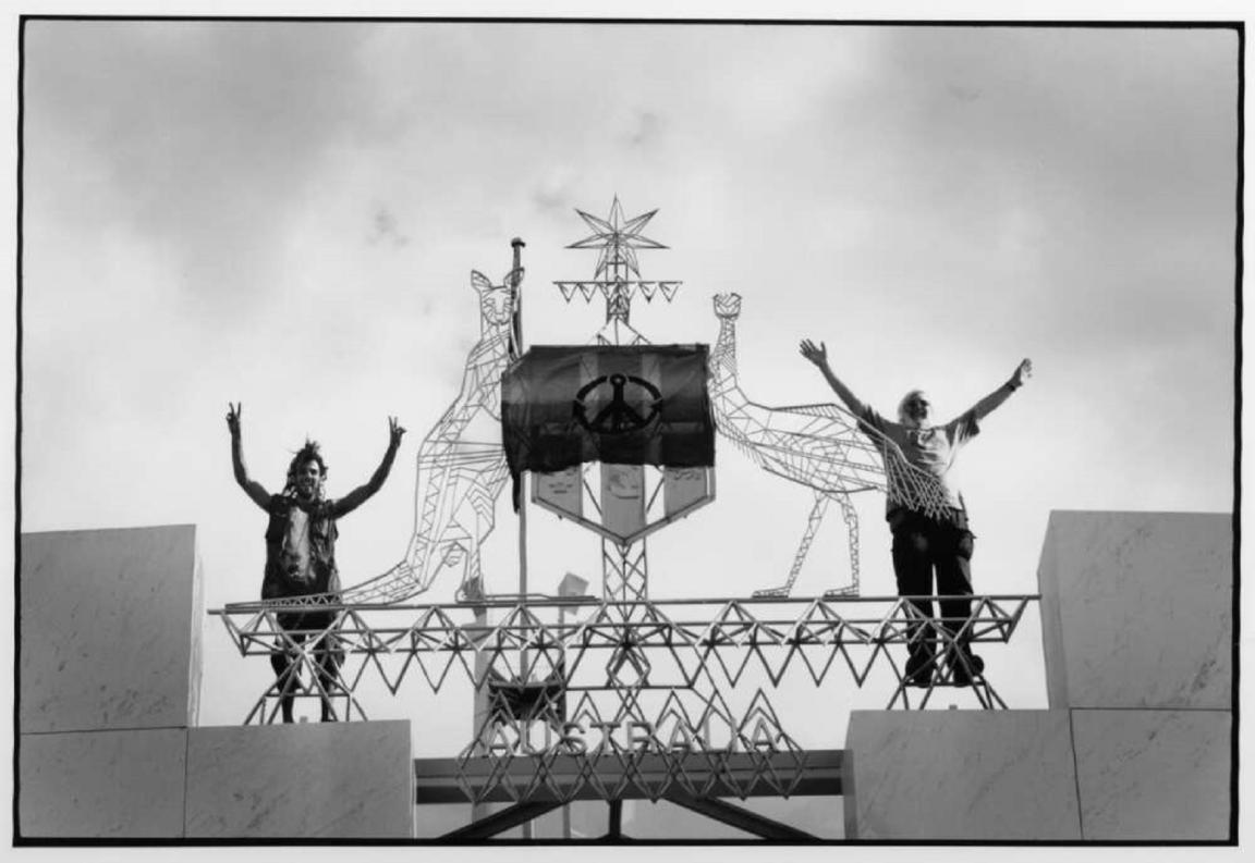 Two demonstrators stand on a structure at Parliament House, Canberra, raising their arms in the air beside an Australian Coat of Arms sculpture. A large peace flag is draped over the emblem. The black-and-white image captures the scene from below, with a cloudy sky in the background.