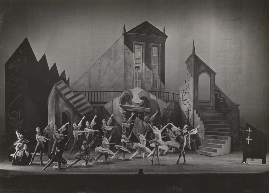  A black-and-white photograph of a ballet performance on stage. The dancers are positioned in elegant formations, some mid-pirouette or in extended poses, wearing costumes that range from tutus to draped garments. The set is an abstract, theatrical backdrop featuring towering, geometric structures, stairs, and archways with a mix of classical and surrealist elements. The lighting creates shadows, adding depth to the scene.