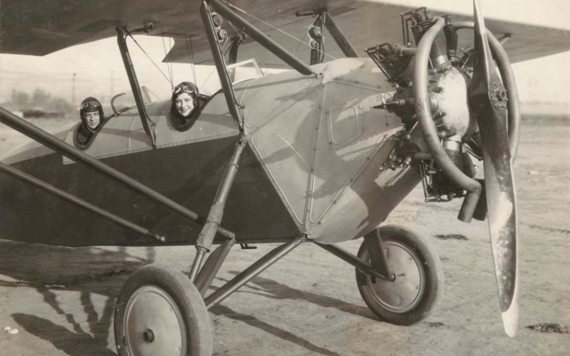 A black and white image of two people sitting in a 1920s style aeroplane while grounded.