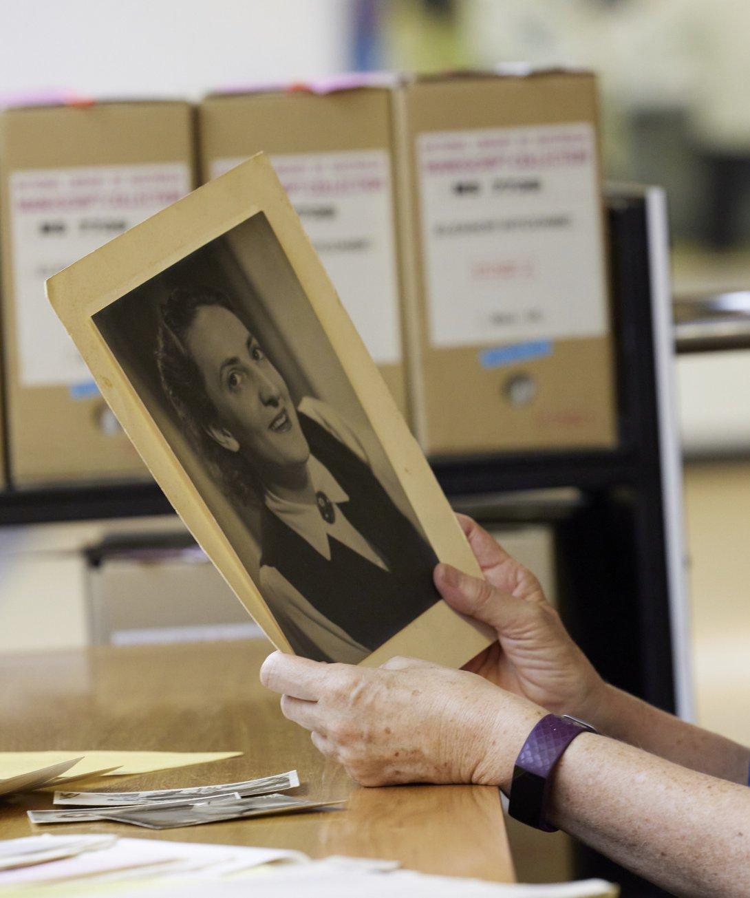 Hands holding up an A4 sized photo of a woman smiling. On the table below are smaller photographs of the same woman and behind is a trolley full of boxes