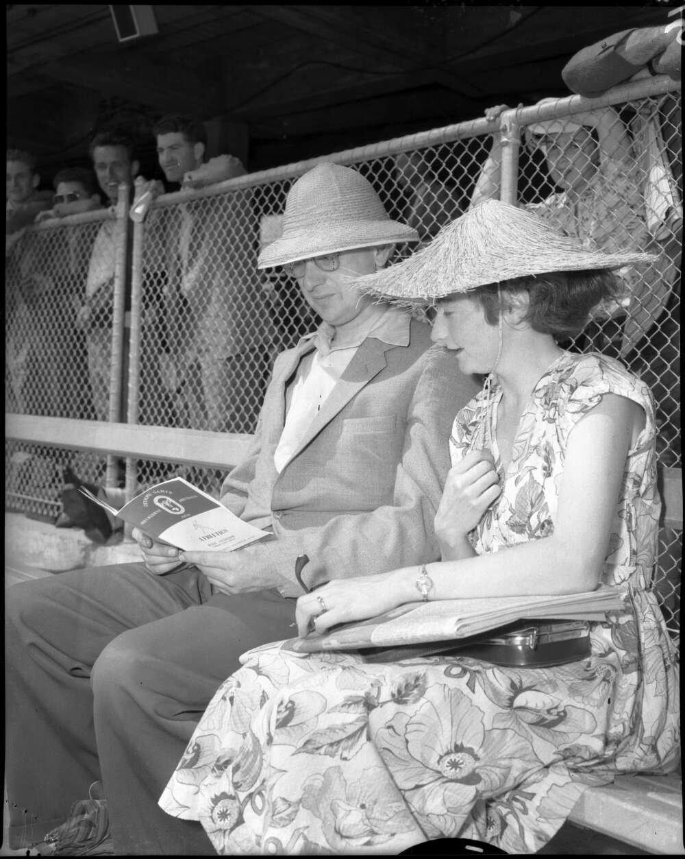 Man and woman sitting in the stands of a sports arena wearing nice clothes and sunhats