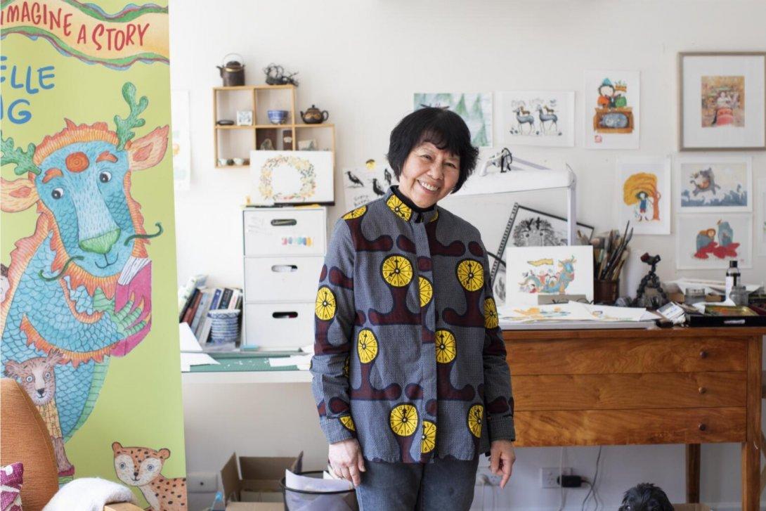 A person standing in the middle of a room. Behind them is a desk covered in illustrations. There are also illustrations on the wall behind the desk. Next to the person is a banner showing a dragon holding a book. The text on the banner reads 'Imagine a story'.