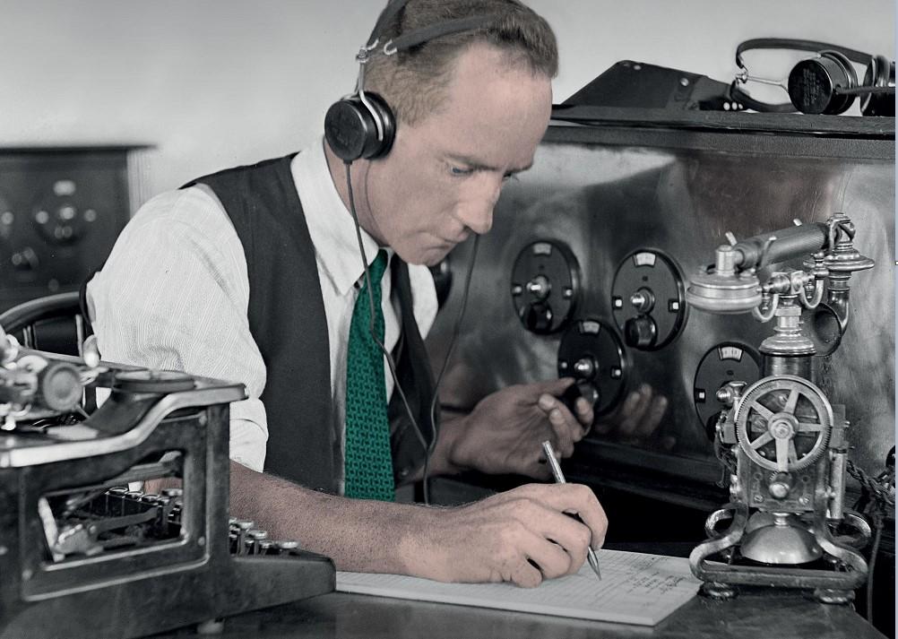 A man wearing old fashioned headphones is sitting at a table. He is writing on a notepad and is surrounded by lots of old fashioned communications equipment.