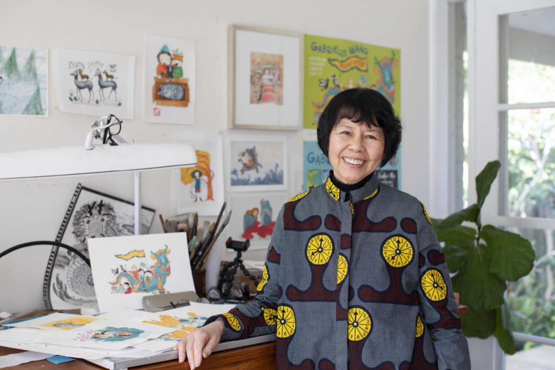 A person leaning against a desk that has illustrations and drawing on it. There are also illustrations and artworks on the wall behind the desk.