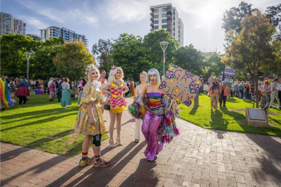 A group of four people who are very colourfully dressed, holding signs in a park. There are more groups of colourful people in the background.