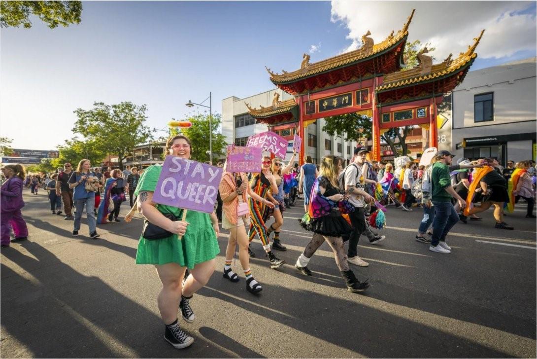 A crowd of people walking along a street next to a Chinese-style gate. The people are holding signs and dressed in colourful clothes.