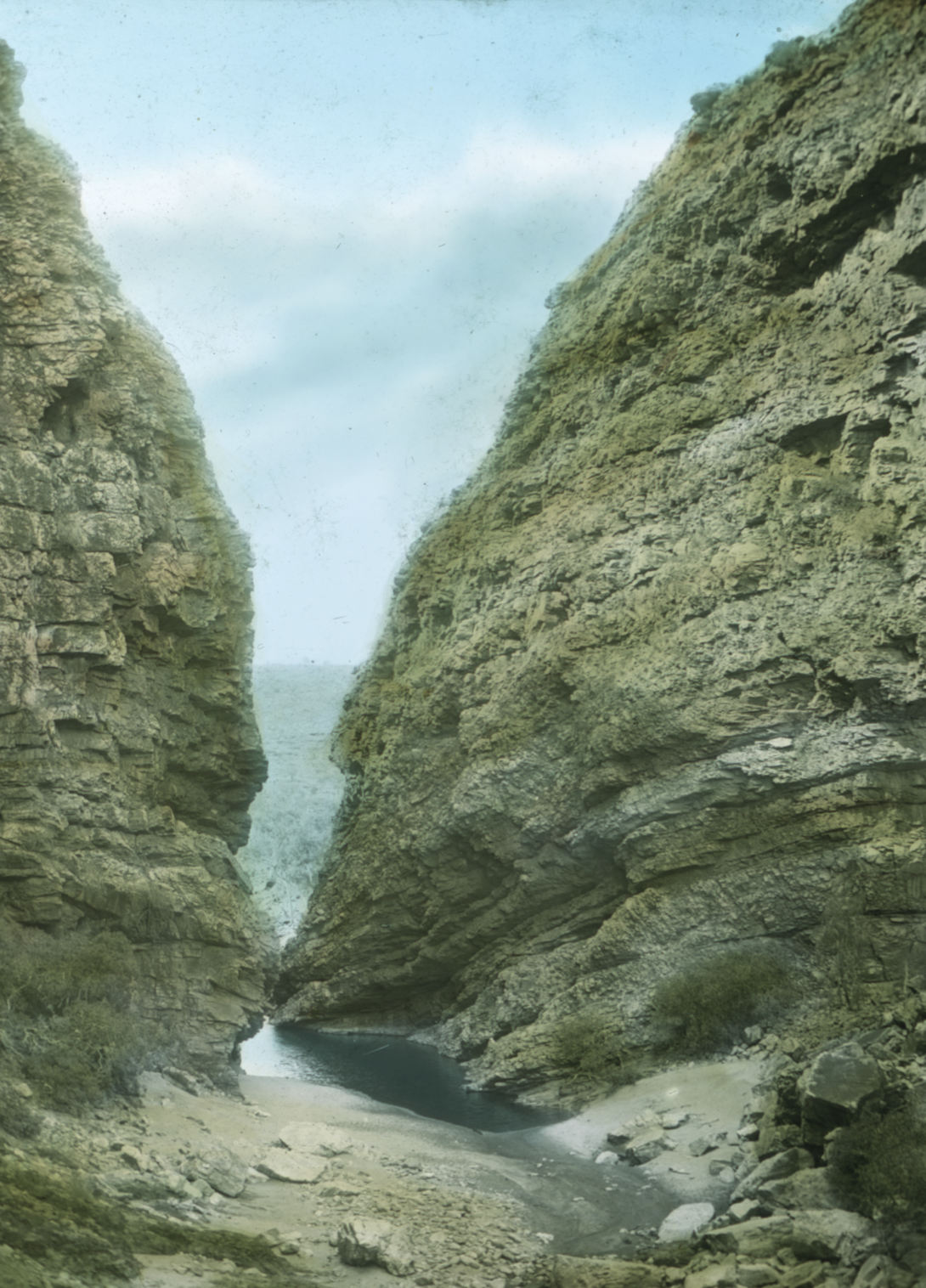 Photo of gap between two cliff-faces. In the gap is a small wateringhole and rocky ground, and a blue sky can be seen