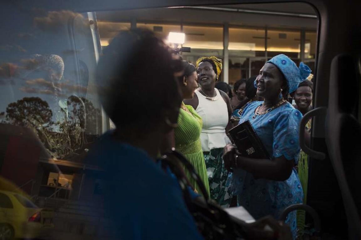 A group of women in colourful traditional clothing stand outside a vehicle, with reflections of trees and the sky visible in the window. One woman, wearing a blue dress and headscarf, holds a small book. The women are gathered closely together, engaging in conversation. The background shows a building with large windows, and the lighting suggests it is late in the day.