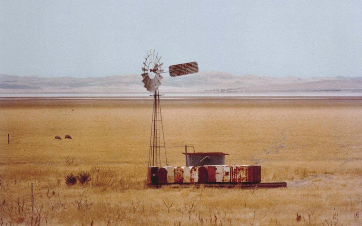 A windmill stands in a paddock surrounded by dry, yellow grass. Lake George in New South Wales is visible in the background.