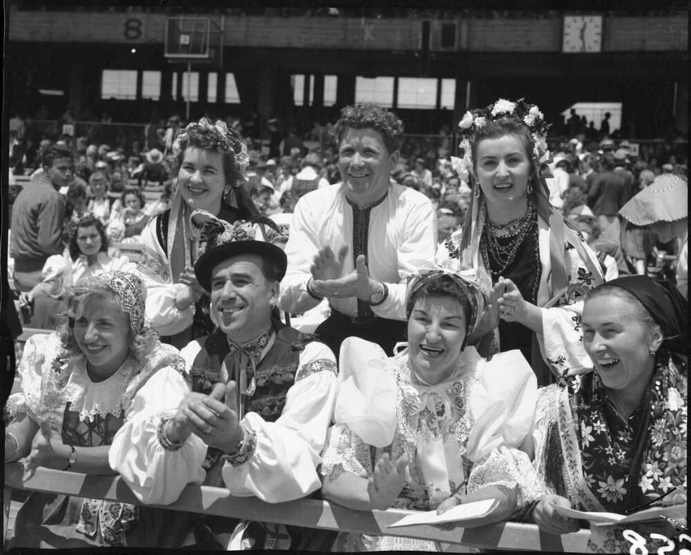 Group of men and women leaning on and standing behind railing as they smile and clap. They are wearing national dress from Europe