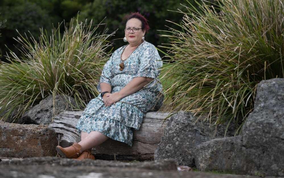 Woman with dark red hair and large round earrings wearing a patterned blue dress and brown ankle boots sitting on a log