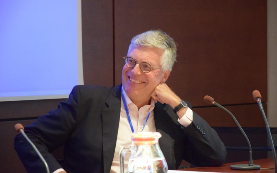 A man sitting at a table behind a microphone, turning his head and smiling.