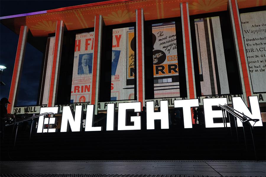 The front of the National Library of Australia, lit up with images and lights. There are big light up letters in front of the building that spell out 'Enlighten'.