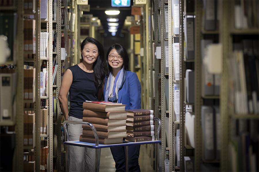 Two women standing behind a trolley with books on it in the Library stacks.