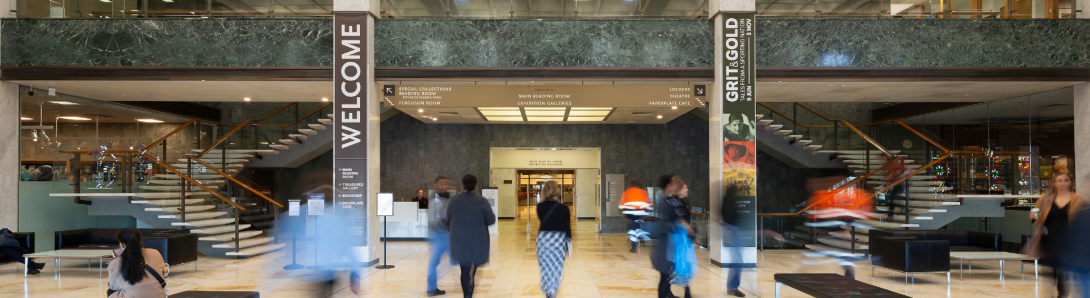 Large two-story room with cream-coloured marble floors, columns and stairs; black leather couches; lots of light from the ceiling and a balcony from the second floor looking over the room. On the columns are signs with directions and the word 'Welcome'. People are moving around the room