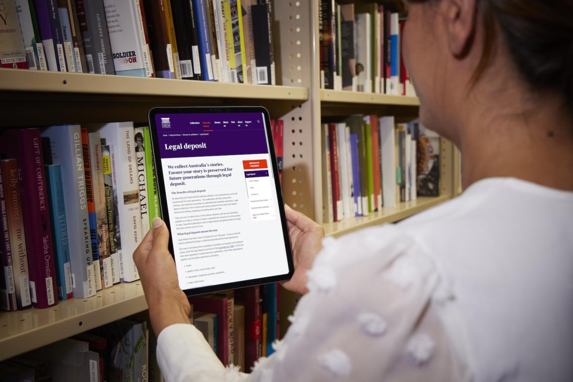 Woman standing in front of a large bookshelf holding up a tablet showing the 'Legal Deposit' page of the National Library website