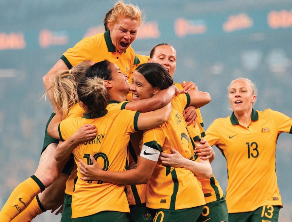 Seven women in the Australian soccer uniform all cheering and hugging one another.