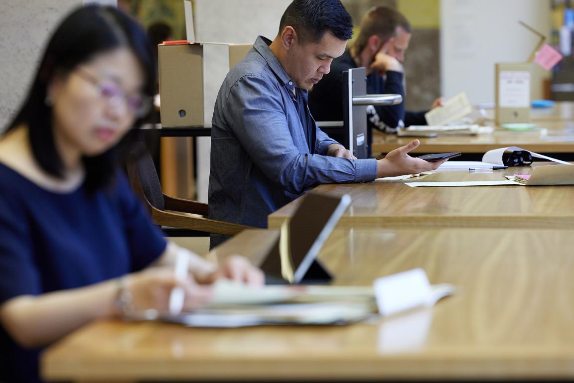 Three people sitting at wooden tables, looking at National Library collection material.