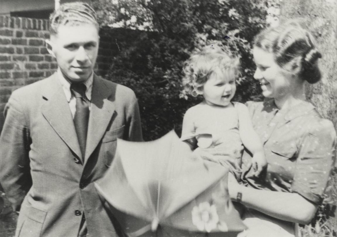 A black and white image of a man standing next to a woman holding a child and an umbrella.