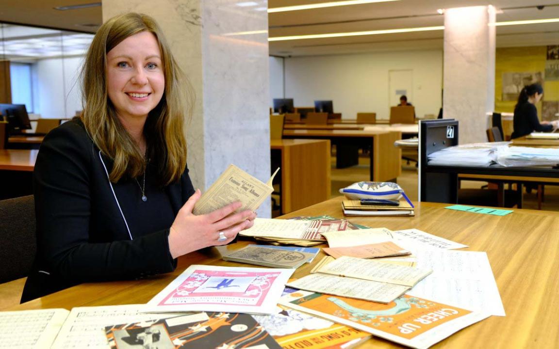 A person with long hair sitting at a table in a big room with lots of papers in front of them.