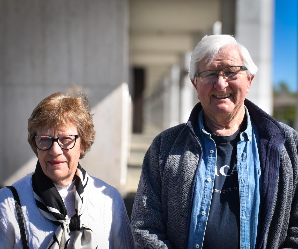 A man and woman, both Library volunteers, standing outside the Library on a sunny day smiling