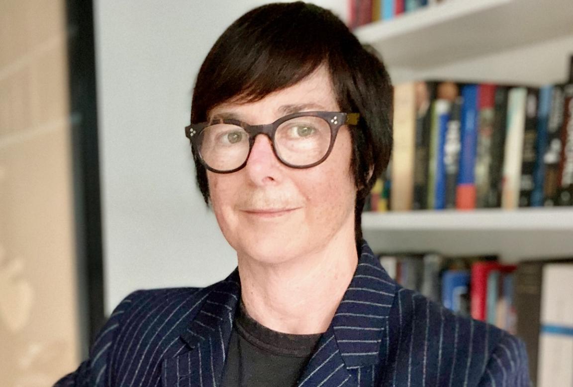 A portrait image of a person with short hair, wearing glasses and a dark blue striped blazer. They are standing in front of a bookshelf.