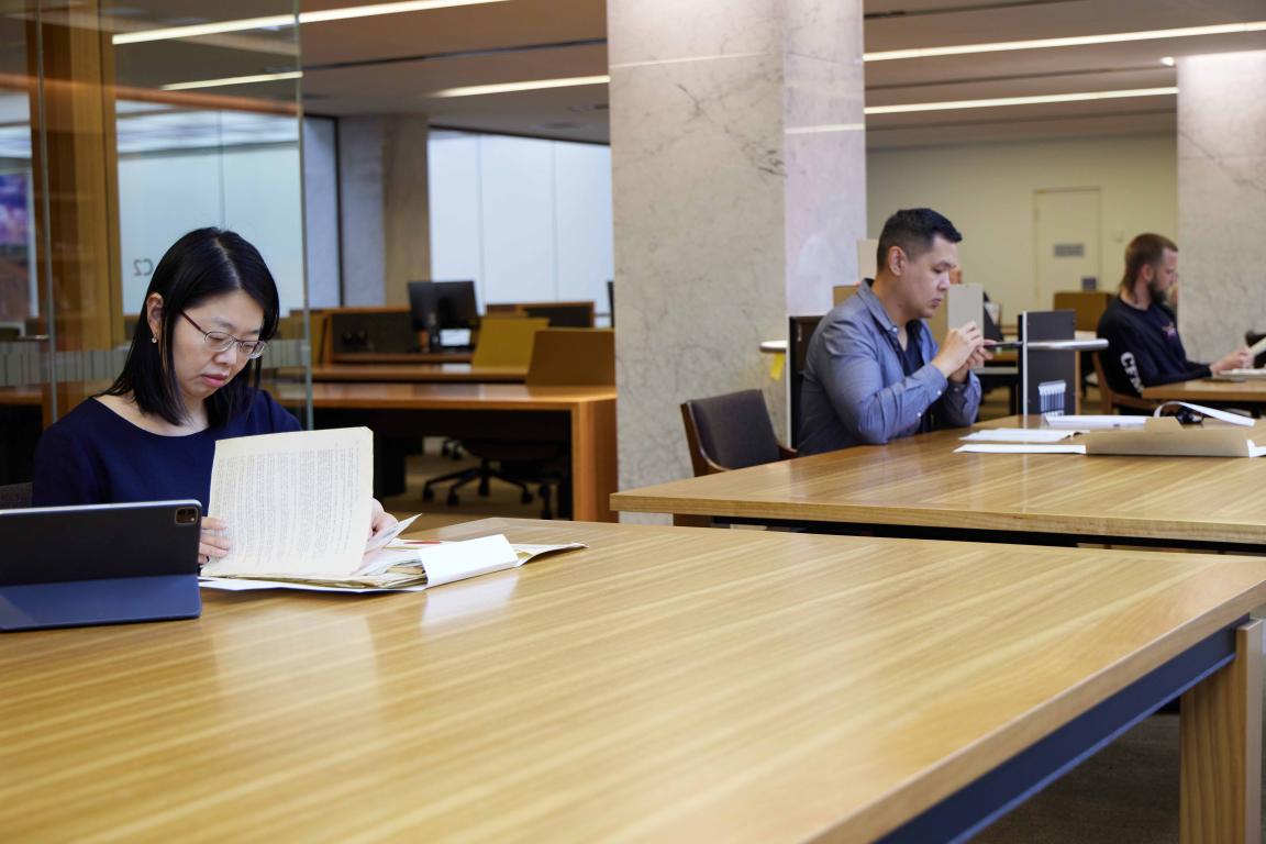 Three people, one woman and two men working at a long table. They are each doing their own research using laptops, notebooks and other resources