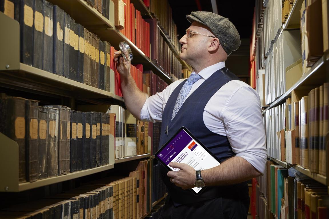 A person standing in front of a bookshelf, holding a magnifying glass up to the shelf. They are also holding an ipad with a website showing on it.