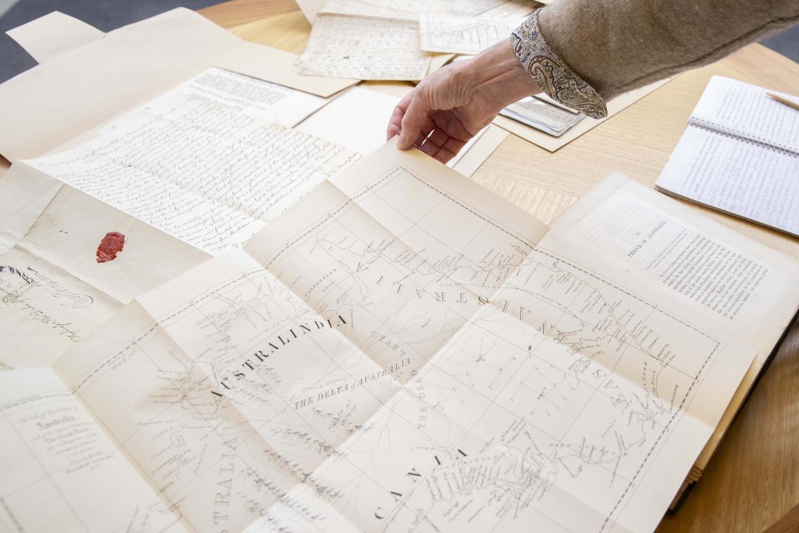 Hand holding up the top corner of a map of Australia, on a table filled with other map and manuscript material